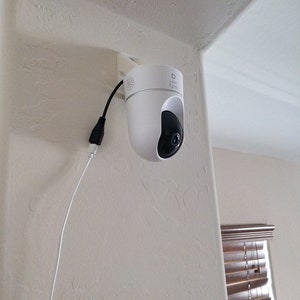Op de afbeelding: Een witte beveiligingscamera met een zwarte lens is gemonteerd op een witte muur. De camera is aangesloten op een wit stroomsnoer.