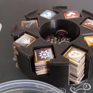 May include: A black plastic organizer with eight compartments for storing small game pieces. Each compartment has a different symbol on it, including a hand, a star, a skull, and a question mark. The organizer has a central cylinder for storing additional pieces.