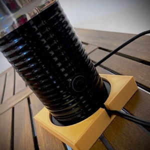 May include: A black and gold charging stand for a coffee maker. The stand has a circular cutout to hold the coffee maker and a slot for the cord. The stand is made of a textured plastic.