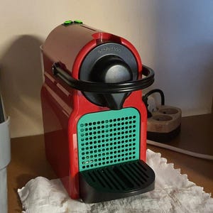 Può includere: Una macchina da caffè Nespresso rossa con la parte superiore nera e un pannello frontale verde. La macchina ha un design elegante e moderno con un corpo curvo e una maniglia nera. Il logo Nespresso è visibile in alto.