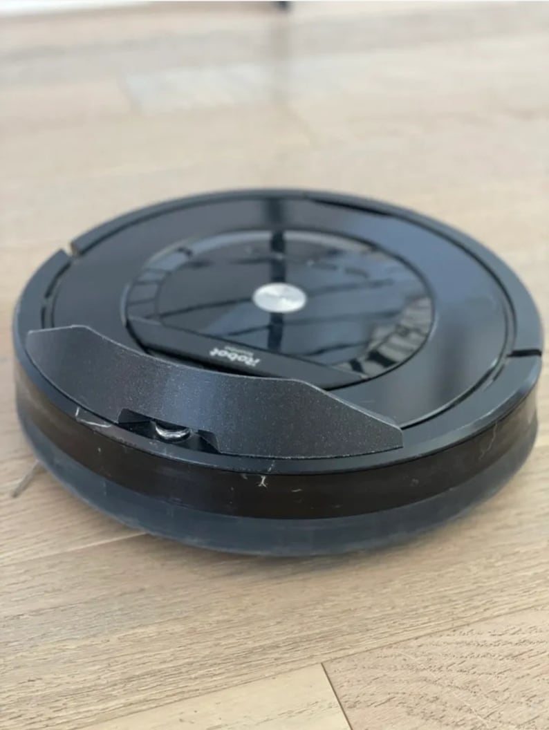 Puede incluir: Un robot aspirador Roomba negro con una parte superior negra y plateada y un parachoques negro. El aspirador est&aacute; en un suelo de madera.