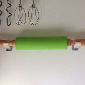 May include: A green silicone rolling pin with wooden handles is mounted on a white wall with two hooks.