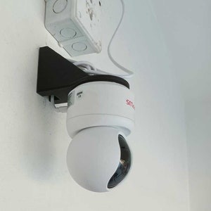 Op de afbeelding: Een witte beveiligingscamera met een zwarte montagebeugel, gemonteerd op een witte muur. De camera heeft een rood "Smart"-logo aan de zijkant.