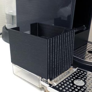 Puede incluir: Máquina de espresso negra con un depósito de agua transparente y una bandeja de goteo plateada. La máquina tiene un portacápsulas incorporado y un exterior negro texturizado. La máquina está sobre una superficie blanca.