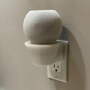 Puede incluir: Soporte de pared blanco impreso en 3D para un altavoz inteligente. El soporte está conectado a una toma de corriente estándar. El altavoz es un dispositivo blanco redondo cubierto de malla.