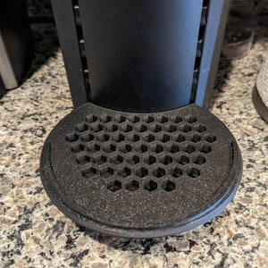 May include: A black coffee maker with a hexagonal drip tray. The drip tray is made of a textured material and has a raised edge.