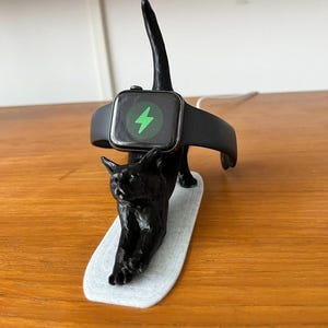 May include: A black cat-shaped charging stand holding a black smartwatch with a green lightning bolt icon on the screen. The stand is on a light gray base, and the watch band is black.