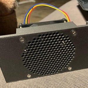May include: A black electronic component with a perforated grill and a fan. The component has a rectangular shape with a series of screws and colorful wires. The item is likely part of a larger electronic device.