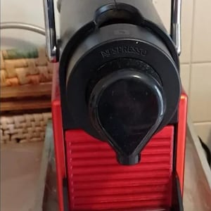 Könnte beinhalten: Eine rot-schwarze Nespresso-Kaffeemaschine mit silbernem Griff. Auf der Vorderseite der Maschine ist der Markenname "Nespresso" aufgedruckt. Sie ist für die Zubereitung von Espresso und anderen Kaffeegetränken konzipiert.