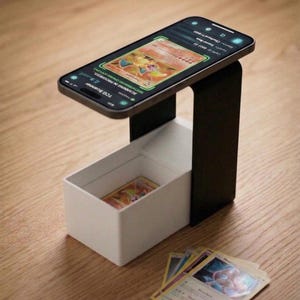 Op de afbeelding: Een zwart-witte telefoonstandaard met een witte opbergdoos. Een smartphone toont een afbeelding van een ruilkaart, met een kaart in de doos en andere in de buurt. Het telefoonscherm toont "TCG Scanner" en kaartdetails.