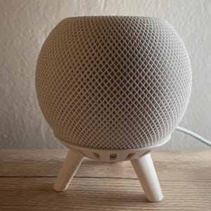 Op de afbeelding: Een witte, ronde slimme speaker met een gaasstofhoes en een driepootstandaard.