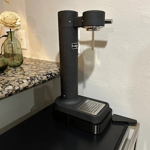 May include: A black countertop soda maker with a silver nozzle and a black base with a metal grate. The soda maker is on a black countertop.