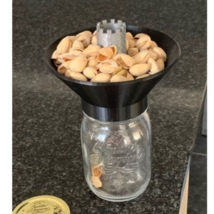May include: A black plastic funnel with a gray plastic top sits on top of a clear glass jar. The funnel is filled with pistachio nuts.