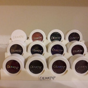 May include: A display of 12 round, white-rimmed, black and purple eyeshadows with the word "COLOURPOP" printed on each. The eyeshadows are arranged in three rows of four on a white shelf with the word "COLOURPOP" printed on the bottom.