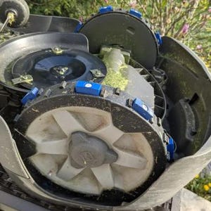 May include: A gray robotic lawnmower with a black and white wheel and blue trim. The lawnmower is partially disassembled, showing the inner workings.