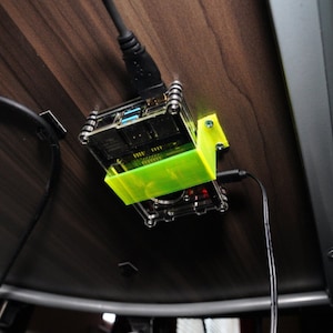 May include: A small black and clear computer case with a green plastic bracket mounted to a wooden surface. The case is connected to a black cable.