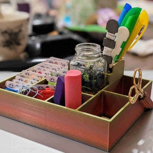 May include: A wooden organizer with multiple compartments for storing craft supplies. It features a clear glass jar, a pink eraser, and a set of colorful pens. The organizer has a red, orange, and yellow gradient finish.