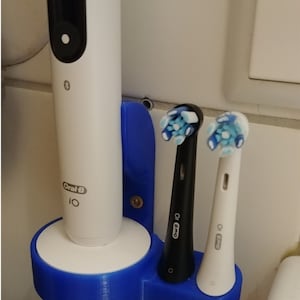 May include: A blue plastic wall-mounted toothbrush holder with three slots. The holder has a white base and a white Oral-B iO toothbrush in the center slot. Two Oral-B Pro electric toothbrush heads are in the side slots.