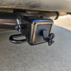 May include: A black metal trailer hitch with a round ball on the end. The hitch is attached to a vehicle.