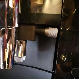 May include: Close-up of a black espresso machine with a silver knob and a chrome spout. The machine has a black and silver finish.