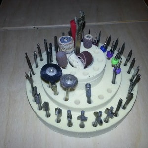 May include: A white plastic rotary tool bit holder with multiple rows of holes for storing bits. The holder is filled with a variety of bits, including sanding drums, grinding stones, and router bits. There are also a few colored rubber bands on some of the bits.