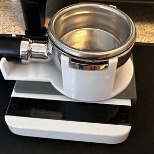 Può includere: Un portafiltro per espresso bianco stampato in 3D con una base grigia, appoggiato su una superficie nera. Il portafiltro è in acciaio inossidabile e ha una finitura cromata.