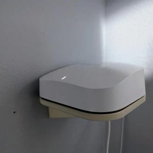 May include: A white, rounded-corner electronic device mounted on a beige bracket. The device has a black trim and a subtle curve on its top surface. A white cable extends from the bottom. The device is mounted on a light gray wall.