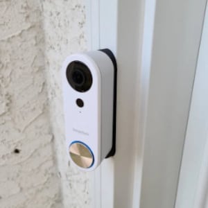 May include: A white doorbell camera with a black mounting plate. The camera has a round lens and a gold button with a blue ring around it.