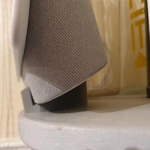 Peut inclure: Un haut-parleur blanc avec une grille en tissu gris et une base noire. Le haut-parleur est posé sur une surface blanche.
