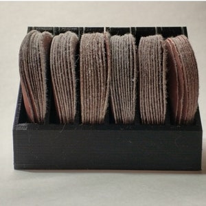 May include: A black plastic storage organizer with six compartments, each holding a stack of round sanding discs. The sanding discs are a light brown color.