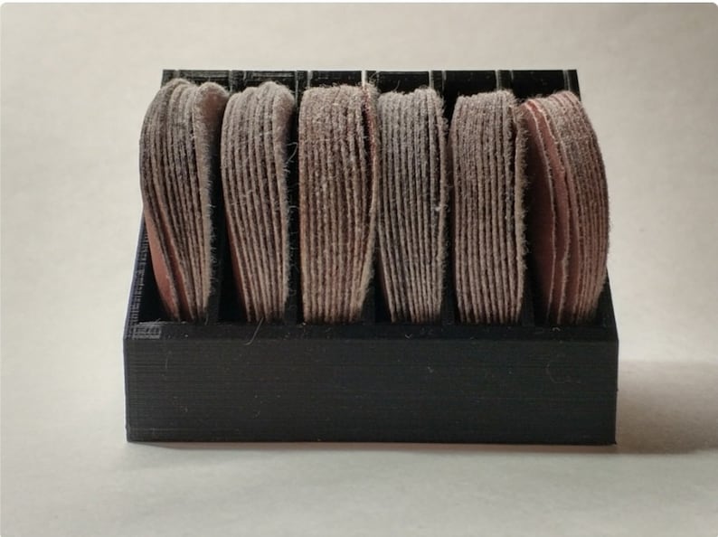 May include: A black plastic storage organizer with six compartments, each holding a stack of round sanding discs. The sanding discs are a light brown color.