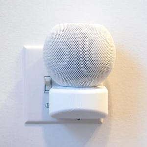 Puede incluir: Un altavoz inteligente blanco con una rejilla de malla está conectado a una toma de corriente blanca.