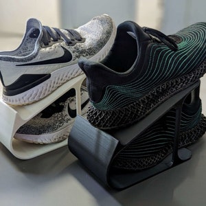 May include: Two pairs of athletic shoes on a white and black shoe rack. The shoes are both black and grey with a white sole. The shoes are on a white shoe rack with a black shoe rack underneath.