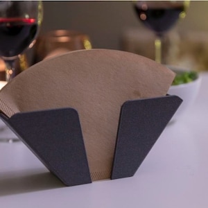 May include: A black metal coffee filter holder with a brown paper filter inside. The holder is shaped like a triangle and has a textured surface.