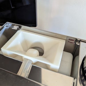 Puede incluir: Primer plano de un embudo de plástico blanco dentro de un electrodoméstico de acero inoxidable. El embudo tiene una pequeña abertura en la parte inferior y una abertura más grande en la parte superior.