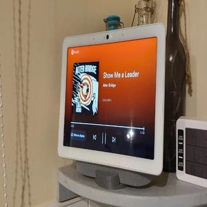 Peut inclure: Un haut-parleur intelligent blanc avec un écran tactile affichant la chanson "Show Me a Leader" d'Alter Bridge sur Spotify. L'écran affiche la pochette de l'album de la chanson, qui présente une image en noir et blanc d'une personne avec une cible sur la tête.