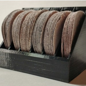 May include: A black plastic storage rack with eight compartments, each holding a brown sanding disc. The sanding discs are stacked on top of each other.