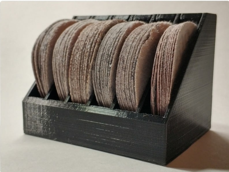 May include: A black plastic storage rack with eight compartments, each holding a brown sanding disc. The sanding discs are stacked on top of each other.