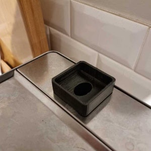 May include: A black plastic square-shaped object with a round hole in the centre. The object is sitting on a silver surface.