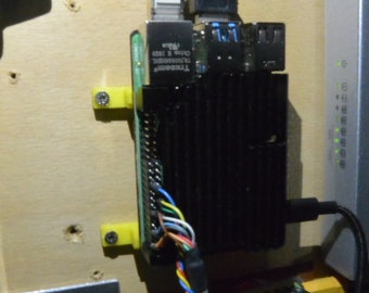 Pode incluir: Um computador Raspberry Pi preto com uma placa de circuito verde e um dissipador de calor preto está montado em uma superfície de madeira com suportes de plástico amarelo. O computador está conectado a um cabo Ethernet branco, um cabo USB preto e uma variedade de outros cabos.