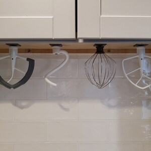 May include: Four different attachments for a stand mixer hang from a wooden rack under a white kitchen cabinet. The attachments include a dough hook, a whisk, a flat beater, and a silicone spatula.