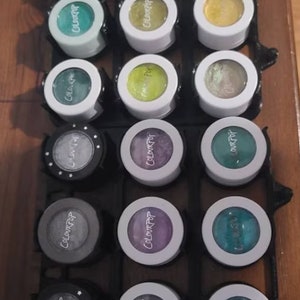May include: A close-up of a set of 12 small round eyeshadow palettes in a black and white organizer. The palettes are arranged in three rows of four. The palettes are all white with black lettering that says "ColourPop". The eyeshadows are various shades of green, blue, purple, and silver.