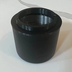 Könnte beinhalten: Ein schwarzer, zylindrischer Behälter aus 3D-gedrucktem Kunststoff mit einem schwarzen Kunststoffring oben. Der Behälter hat eine glatte Oberfläche und der Ring eine strukturierte Oberfläche.