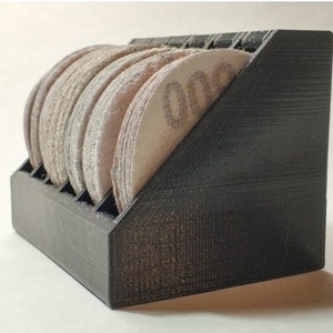 May include: A black 3D printed storage rack for sanding discs. The rack has slots for holding the discs and is angled to allow for easy access. The rack is filled with sanding discs of various grits.