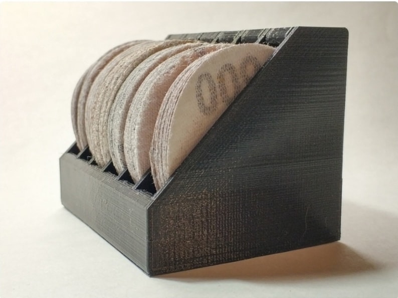 May include: A black 3D printed storage rack for sanding discs. The rack has slots for holding the discs and is angled to allow for easy access. The rack is filled with sanding discs of various grits.