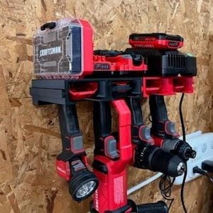Peut inclure: Un organisateur mural pour outils électriques, de couleur rouge et noire. Il peut contenir trois perceuses sans fil et un chargeur de batterie, avec une boîte de rangement en plastique transparent portant l'inscription "CRAFTSMAN" sur le dessus.