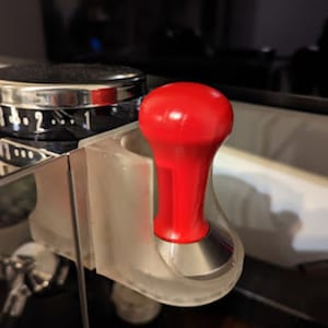 May include: A red and silver espresso tamper with a clear plastic holder. The tamper is attached to the holder with a silver screw.