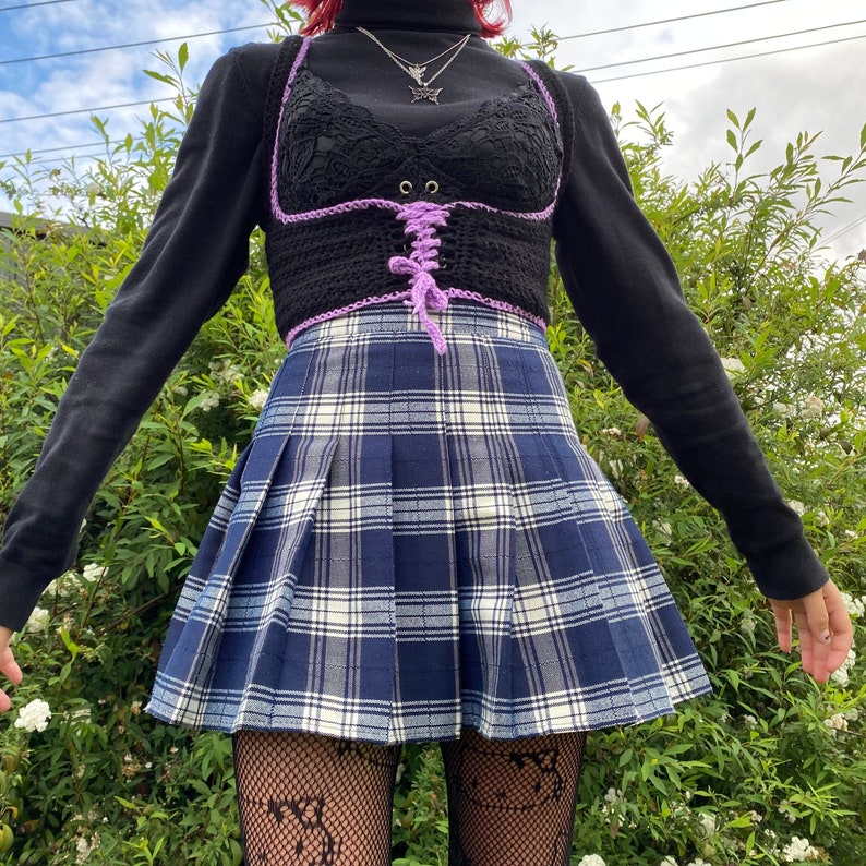 K&ouml;nnte beinhalten: Ein schwarzes Langarmshirt mit einem lila und schwarzen H&auml;keltop und einem blau-wei&szlig; karierten Faltenrock.