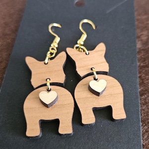 May include: A pair of wooden earrings shaped like a dog's rear end with a small heart charm hanging from each. The earrings are a light brown color and have gold-colored hooks.