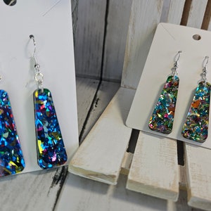 May include: Two pairs of dangle earrings with silver hooks. The earrings are made of clear resin with iridescent glitter embedded in the resin. One pair is blue and the other is green.